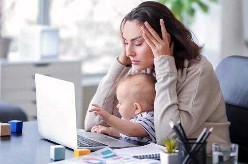 Майка с дете на ръце, работеща на лаптоп у дома.0
