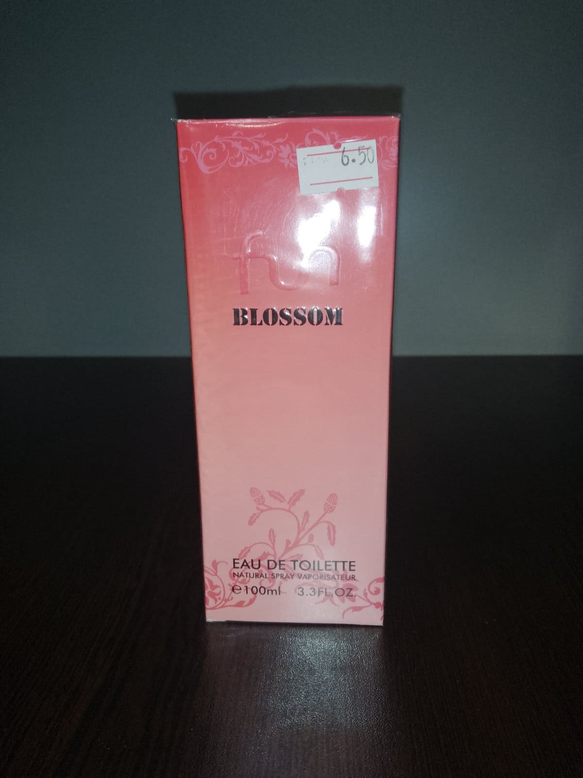 Флакон на тоалетна вода Fun Blossom с розов дизайн и златиста капачка, поставен до розова опаковка върху дървена повърхност.1