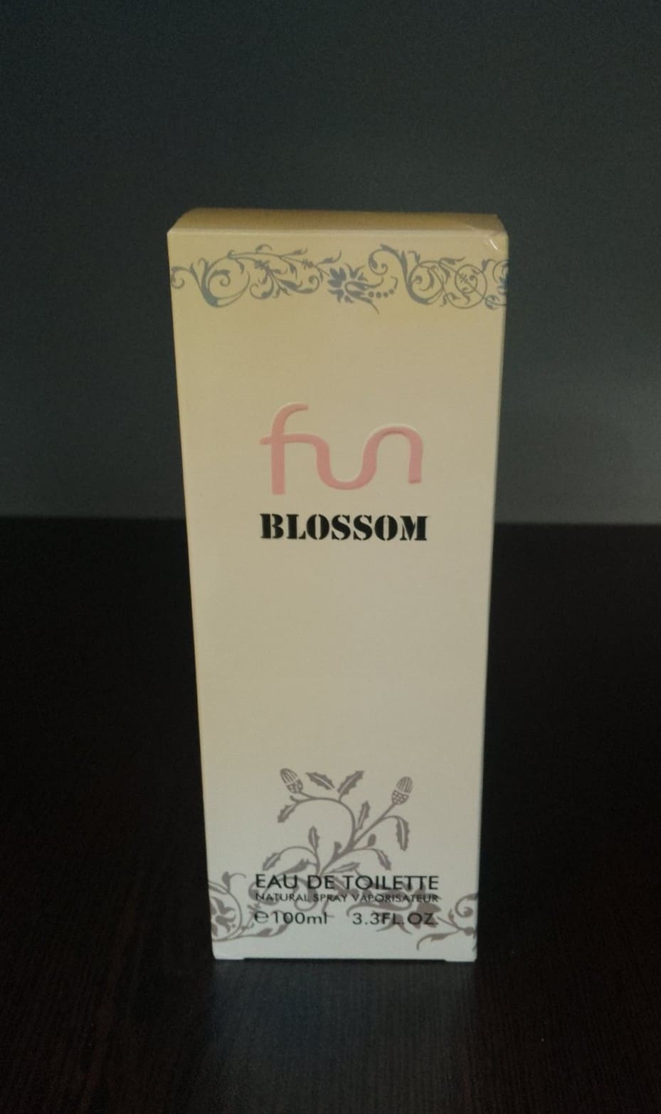 Флакон на тоалетна вода Fun Blossom със златиста капачка и прозрачен дизайн, до светла елегантна опаковка върху дървена повърхност.1